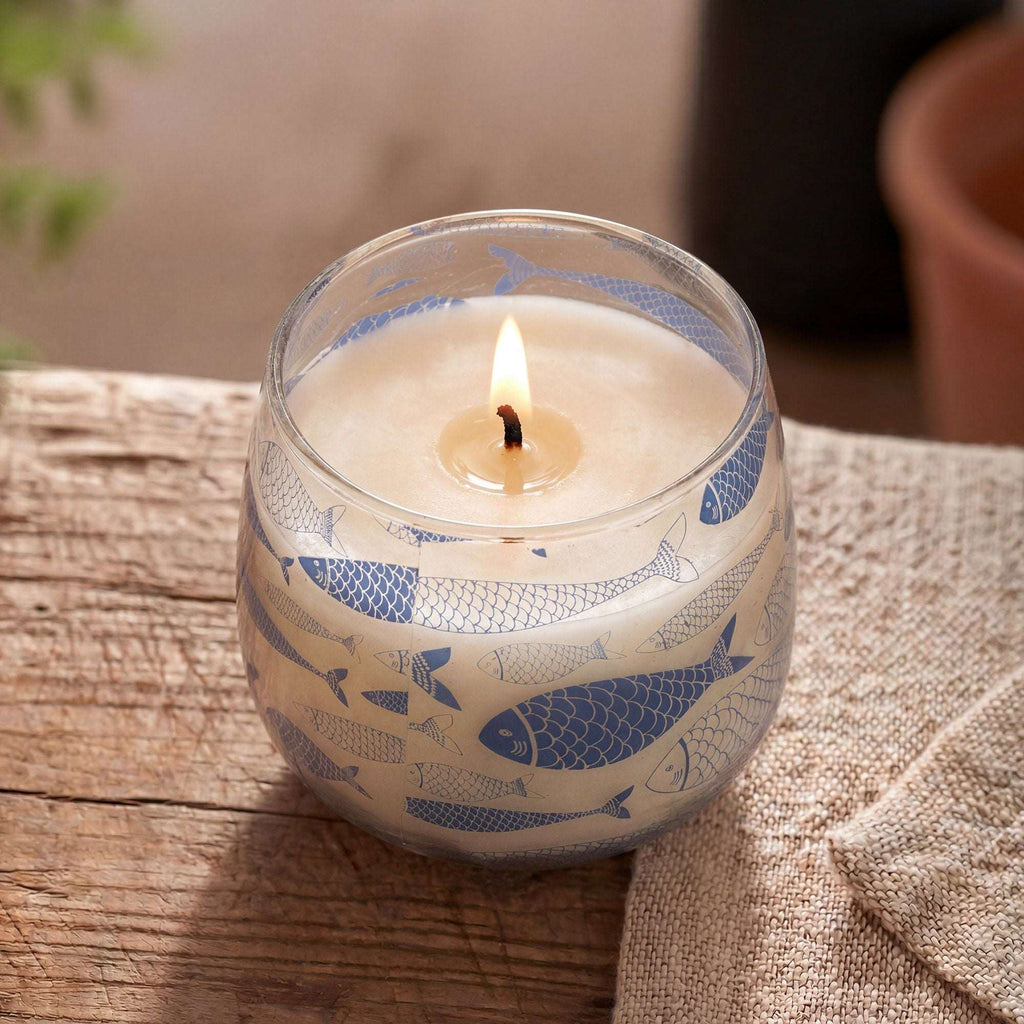 Medium Glass Jar Candles - Seabreeze with a lit wick inside a clear glass jar decorated with blue fish patterns, set on a rustic wooden surface.