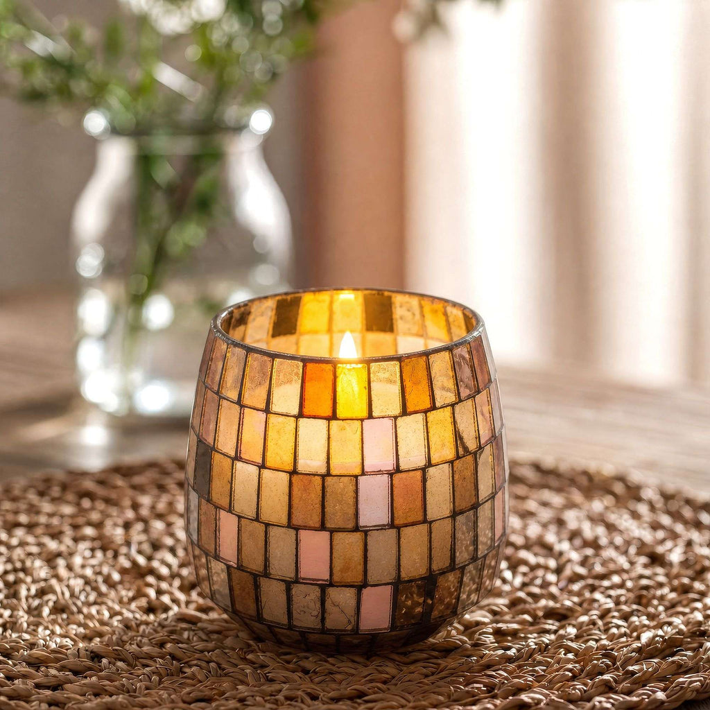 Medium Glass Jar Candles - Amber Mosaic featuring warm amber and cream mosaic glass pieces, softly glowing with a lit candle inside, placed on a woven surface with blurred background elements.