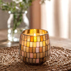 Medium Glass Jar Candles - Amber Mosaic featuring warm amber and cream mosaic glass pieces, softly glowing with a lit candle inside, placed on a woven surface with blurred background elements.