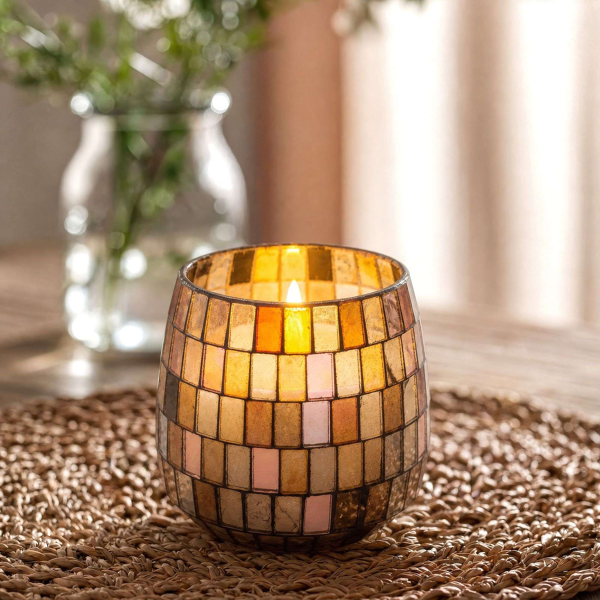 Medium Glass Jar Candles - Amber Mosaic featuring warm amber and cream mosaic glass pieces, softly glowing with a lit candle inside, placed on a woven surface with blurred background elements.