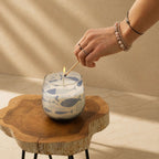 A hand with delicate bracelets lights a wick of a Medium Glass Jar Candles - Seabreeze featuring blue fish patterns, sitting on a rustic wooden stool in soft natural light.