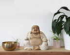 Happy Buddha - Whitewash 30cm sitting statue with a serene smile and prayer beads, displayed on a wooden surface alongside potted plants and a brass bowl, blending natural and spiritual decor.