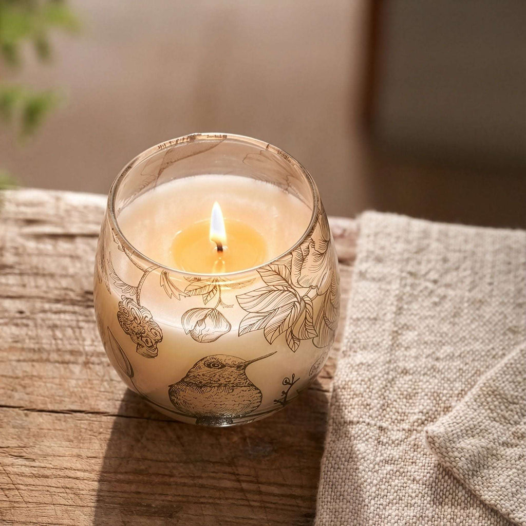 Lit candle in a small glass jar decorated with delicate botanical and bird illustrations, named Small Glass Jar Candles - Drawn by Flame, placed on a rustic wooden surface next to a beige textile.