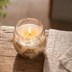 Lit candle in a small glass jar decorated with delicate botanical and bird illustrations, named Small Glass Jar Candles - Drawn by Flame, placed on a rustic wooden surface next to a beige textile.