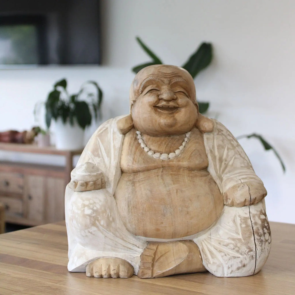 Happy Buddha - Whitewash 30cm sitting smiling with a beaded necklace and a smooth whitewashed carved finish, displayed on a wooden surface with indoor plants in the background.