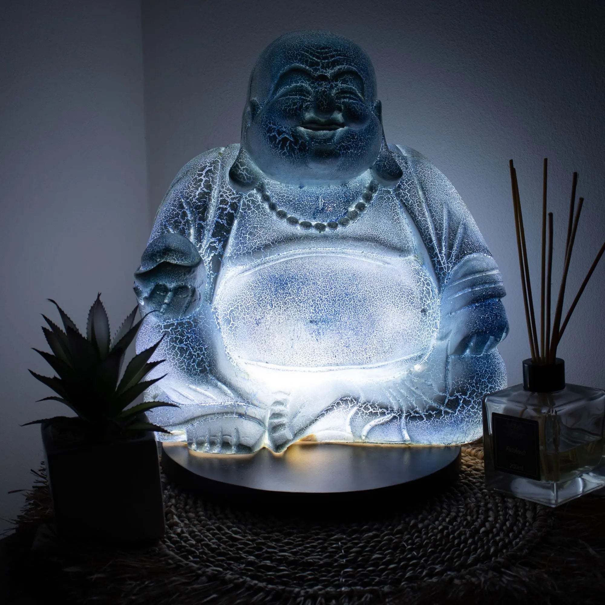 Laughing Buddha Lamp Sky Blue - Round Stand glowing softly with textured resin surface, creating a peaceful ambiance alongside decorative plants and reed diffuser.