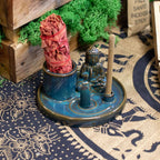 Multi Purpose Buddha Incense Plate - Ceramic Blue with a small Buddha statue figurine, holding a blue incense cone and a stick, placed on a patterned cloth with green moss in the background.
