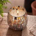 Lit Medium Glass Jar Candles - Amber Mosaic with a mosaic glass pattern in warm amber and neutral tones sitting on a wooden surface next to a beige cloth.