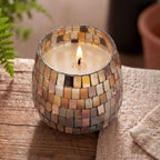 Lit Medium Glass Jar Candles - Amber Mosaic with a mosaic glass pattern in warm amber and neutral tones sitting on a wooden surface next to a beige cloth.