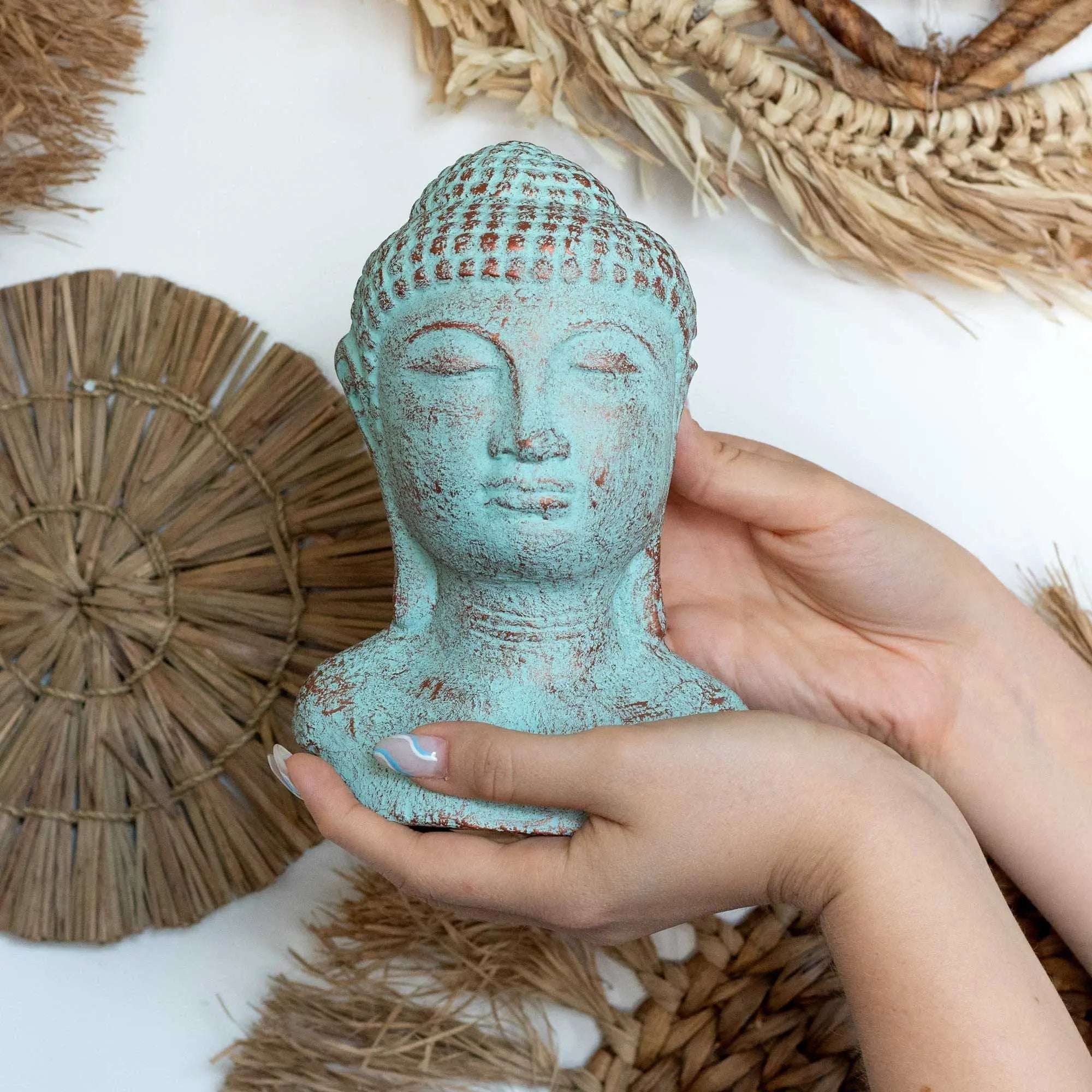 Hands gently holding a Tranquility Buddha Head - Green Copper with a peaceful expression, surrounded by natural woven decor elements for serene Buddha decor.