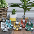 A collection of Silver Mini Buddha (Assorted Colours) statues in silver, turquoise, gold, orange, and green finishes, arranged on a wooden surface with potted plants in the background.