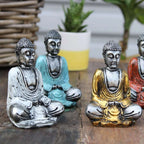 Four Silver Mini Buddha (Assorted Colours) statues in silver, turquoise, gold, and red seated in meditation on a wooden surface with a woven basket and potted plant in the background.