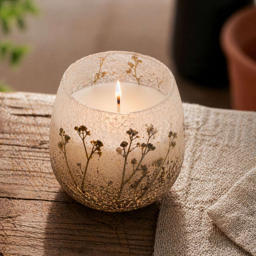 Medium Glass Jar Candles - Wildlight Woods featuring a lit candle in a textured glass jar with botanical details, placed on a rustic wooden surface with a neutral fabric nearby.