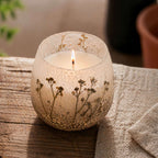 Medium Glass Jar Candles - Wildlight Woods featuring a lit candle in a textured glass jar with botanical details, placed on a rustic wooden surface with a neutral fabric nearby.