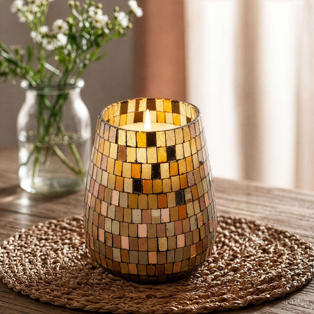 Lit Tall Glass Jar Candles - Amber Mosaic with warm amber and gold mosaic tiles resting on a woven mat, softly glowing in a cozy indoor setting with blurred floral background.