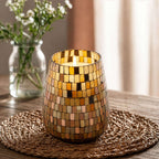 Lit Tall Glass Jar Candles - Amber Mosaic with warm amber and gold mosaic tiles resting on a woven mat, softly glowing in a cozy indoor setting with blurred floral background.