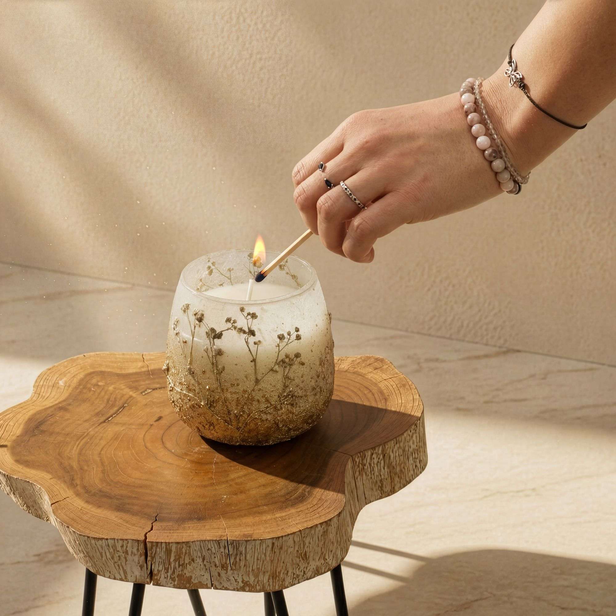 Hand lighting a Medium Glass Jar Candles - Wildlight Woods on a natural wood table, showcasing its frosted glass with embedded dried floral accents.
