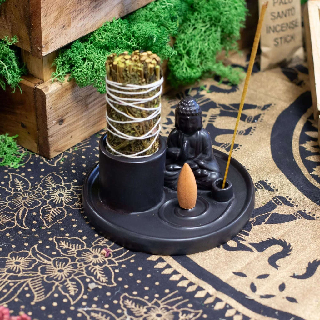 Multi Purpose Buddha Incense Plate - Ceramic Matt Black featuring a seated Buddha statue, with compartments holding incense sticks, cones, and a bundle of sage, set on a patterned cloth.