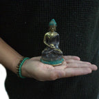 Person holding a small Med Meditation Sitting Buddha statue in their open hand, featuring a dark and gold resin finish with a turquoise base and serene facial expression.