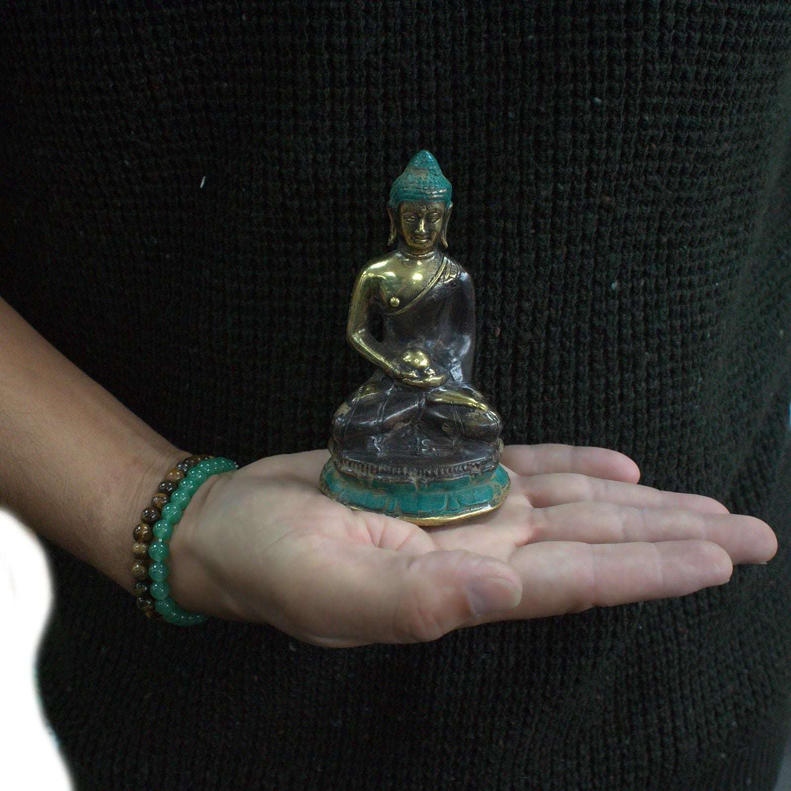 Person holding a small Med Meditation Sitting Buddha statue in their open hand, featuring a dark and gold resin finish with a turquoise base and serene facial expression.
