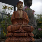 Hand Carved Buddha Statue - 80cm Welcome seated in meditation with hands in prayer, featuring detailed wood carving and a serene expression surrounded by garden plants.