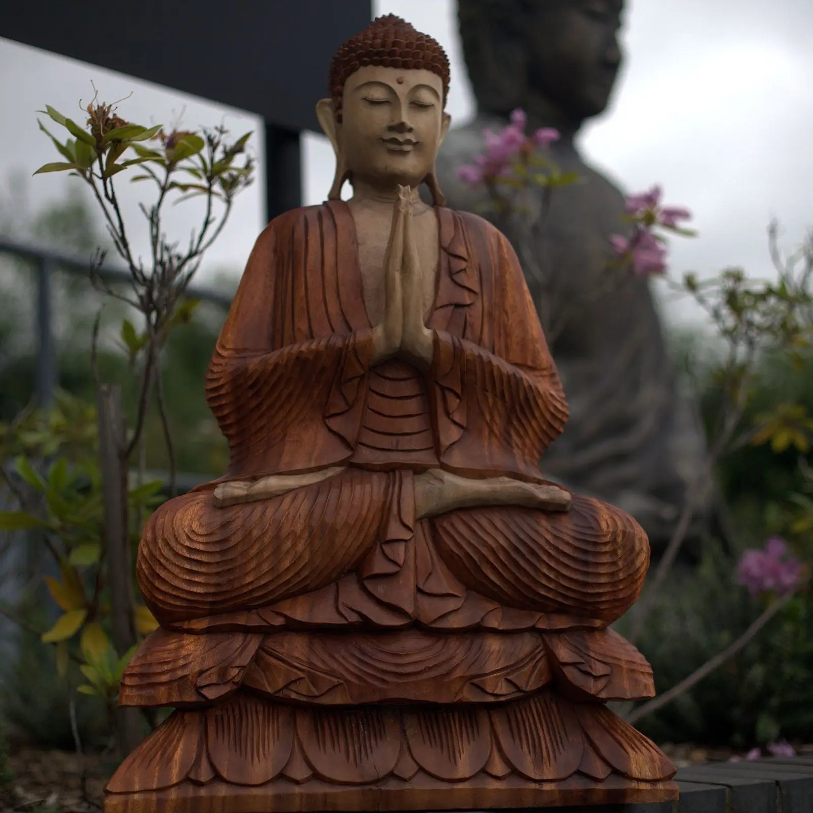 Hand Carved Buddha Statue - 80cm Welcome showing a detailed wooden Buddha in a peaceful meditative pose with hands in prayer, surrounded by greenery and flowers.