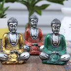 Three Silver Mini Buddha (Assorted Colours) statues in yellow, red, and green sitting in meditation pose on a wooden surface with blurred plants in the background.