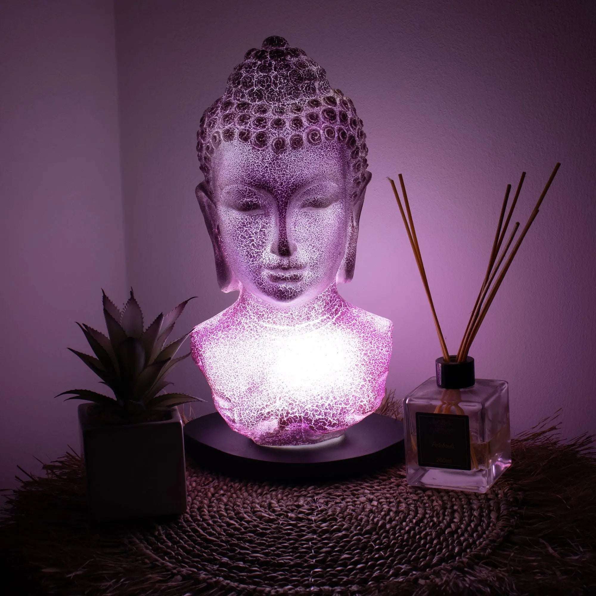 Buddha Bust Lamp Lavender Crackle - Oval Stand glowing softly in lavender light, placed on a woven mat beside a plant and reed diffuser, creating a serene ambiance.