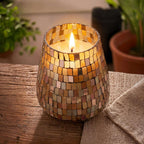 Lit candle in a tall glass jar covered with amber mosaic tiles, placed on a wooden surface next to a beige cloth and surrounded by blurred green plants, showcasing Tall Glass Jar Candles - Amber Mosaic.