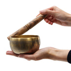 Hands gently holding and playing a Five Buddha Singing Bowl with a wooden mallet, showcasing its brass material and engraved design for meditation and mindfulness practices.
