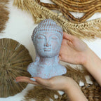 Hands gently holding a Tranquility Buddha Head - Blue Copper statue with a textured, serene face and rustic blue copper finish against a natural woven backdrop.