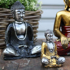 Two Silver Mini Buddha (Assorted Colours) statues featuring detailed serene faces and robes, one in silver and black, the other in silver and gold, displayed on a rustic wooden surface.
