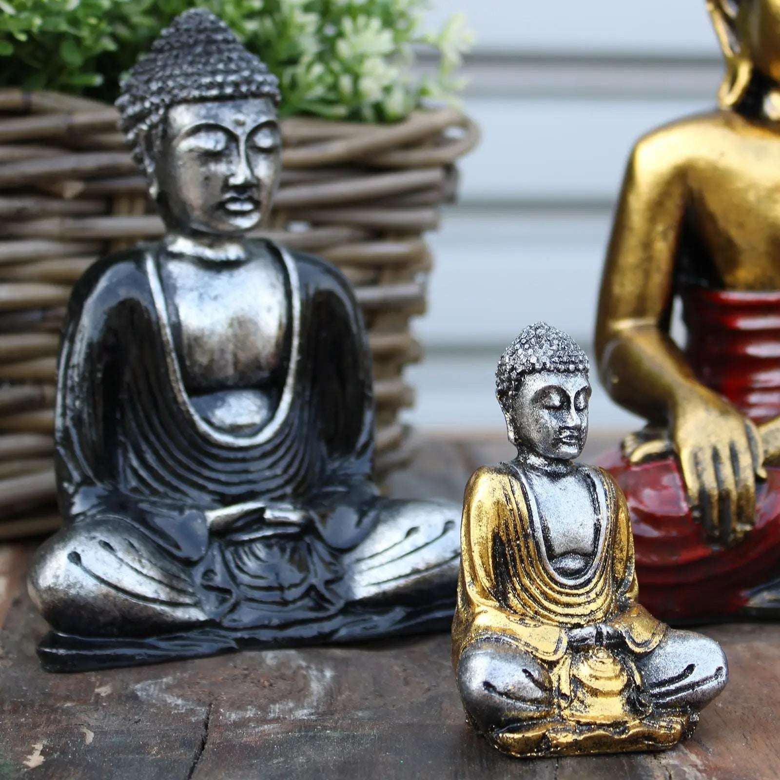 Two Silver Mini Buddha (Assorted Colours) statues featuring detailed serene faces and robes, one in silver and black, the other in silver and gold, displayed on a rustic wooden surface.