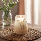A softly glowing Tall Glass Jar Candles - Wildlight Woods with delicate dried flowers inside, placed on a woven mat on a wooden table with a blurred vase of white flowers in the background.