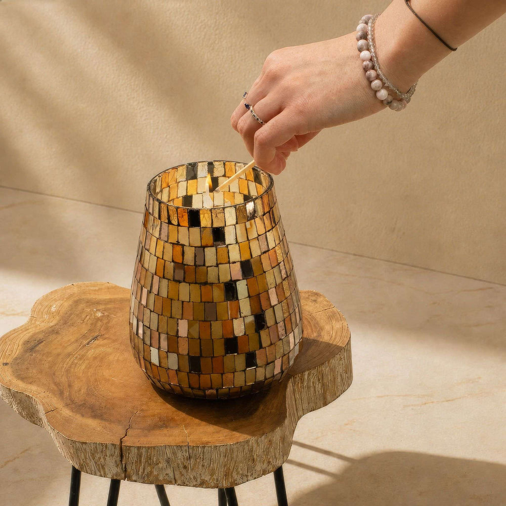 Hand lighting the wick of a Tall Glass Jar Candles - Amber Mosaic with amber, gold, and brown mosaic tiles on a wooden table in warm natural light.