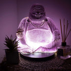 Laughing Buddha Lamp Lavender Crackle - Round Stand glowing with soft purple light, surrounded by decorative plants and a reed diffuser, creating a serene ambiance.