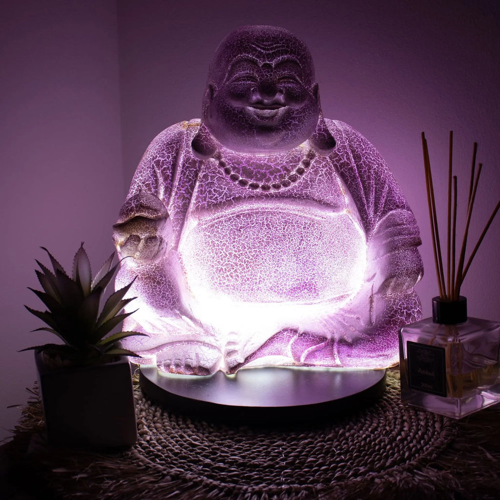 Laughing Buddha Lamp Lavender Crackle - Round Stand glowing softly with a purple crackle texture, placed on a woven mat beside a small plant and reed diffuser.