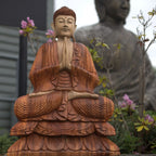 Hand Carved Buddha Statue - 80cm Welcome showing a detailed wooden Buddha figure seated in meditation with hands in prayer position, surrounded by garden plants and flowers.