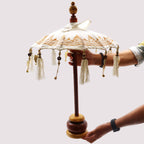 Person holding a Bali Home Decor Parasol - Canvas - Gold Decor- 40cm with cream canvas, gold decorative patterns, wooden handle, and hanging tassels against a plain background.