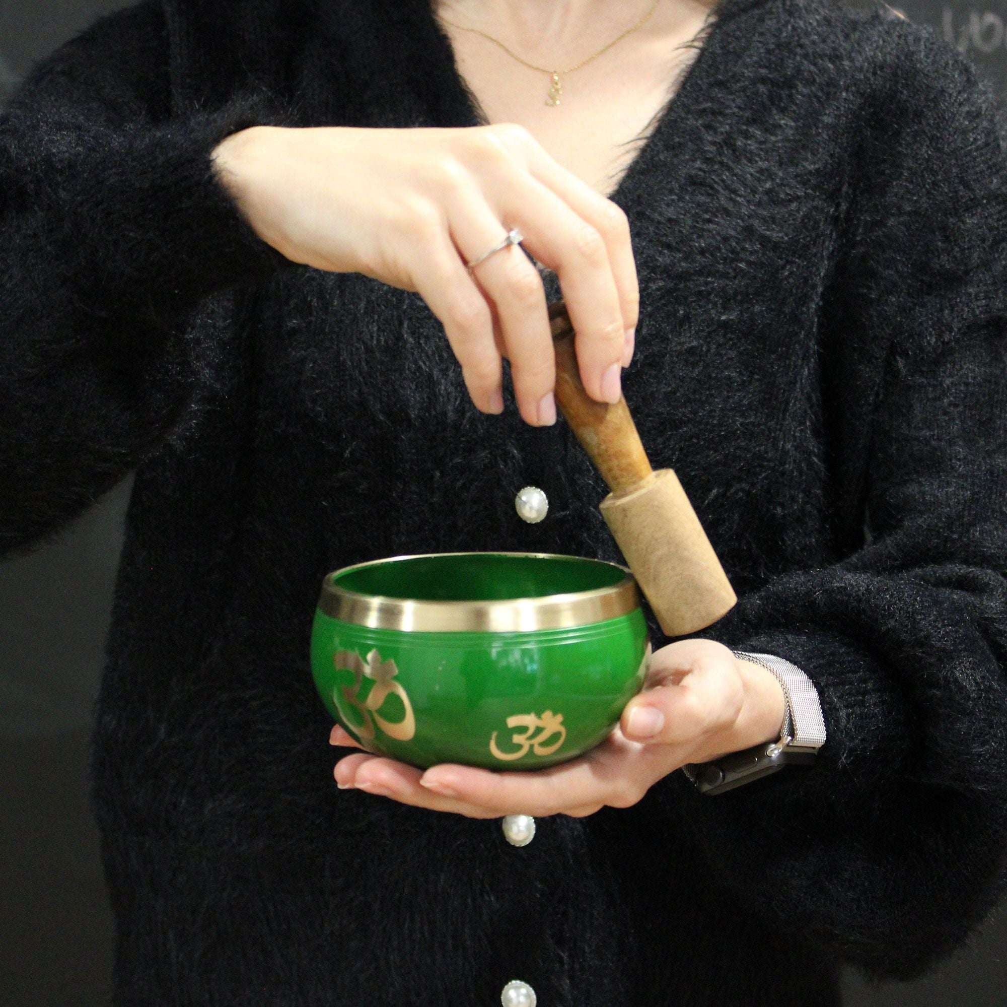 Person holding a green singing bowl with gold Om symbols, using a wooden striker, representing Buddha Singing Bowl Set- Black/Orange 10.7cm.