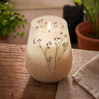 Tall Glass Jar Candles - Wildlight Woods with a lit wick, featuring delicate pressed wildflowers embedded in frosted glass, resting on a rustic wooden surface next to a cozy fabric cloth.