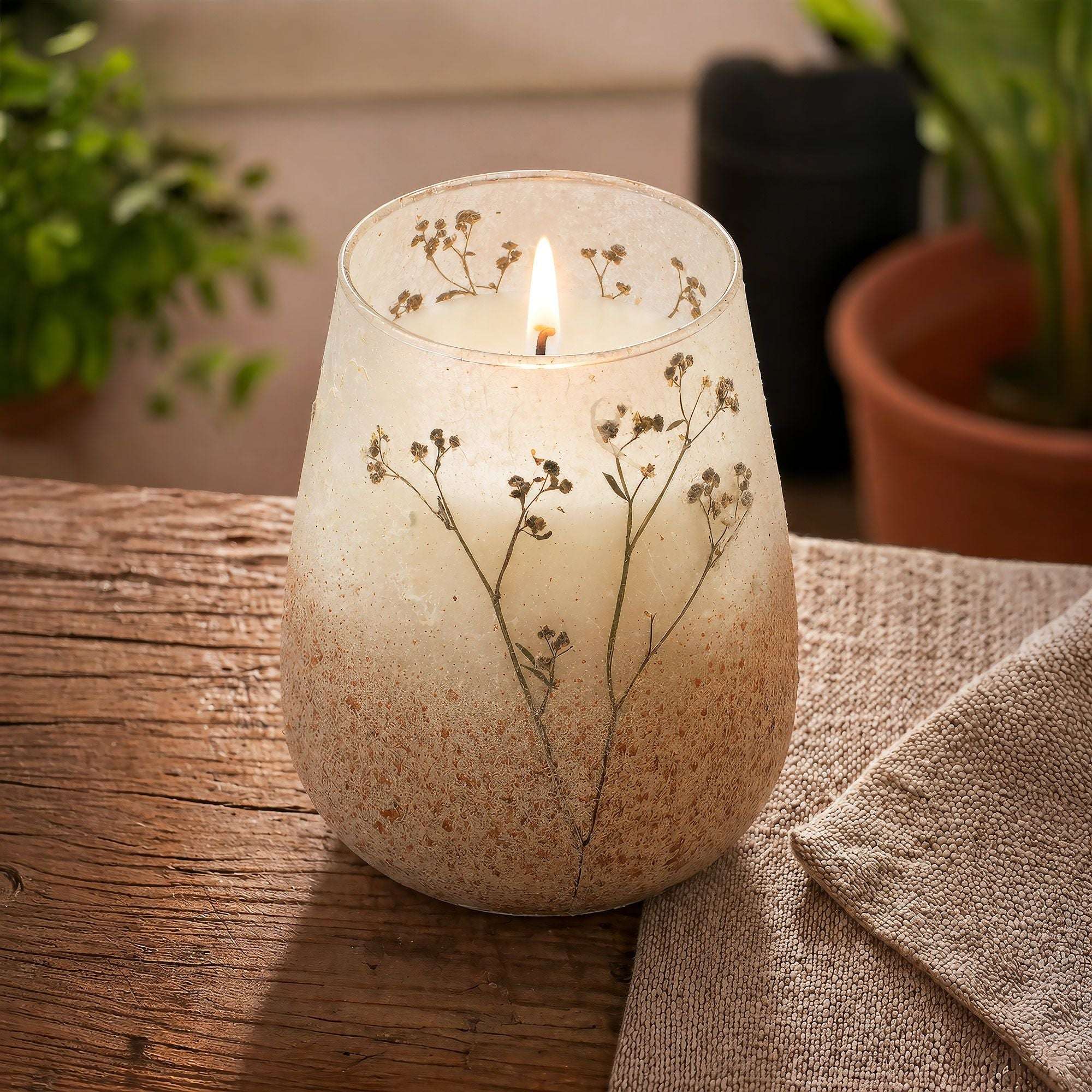 Tall Glass Jar Candles - Wildlight Woods with a lit wick, featuring delicate pressed wildflowers embedded in frosted glass, resting on a rustic wooden surface next to a cozy fabric cloth.
