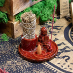 Multi Purpose Buddha Incense Plate - Ceramic Soft Red with seated Buddha figure, holding a bundle of herbs and an incense cone and stick holder, resting on patterned fabric.