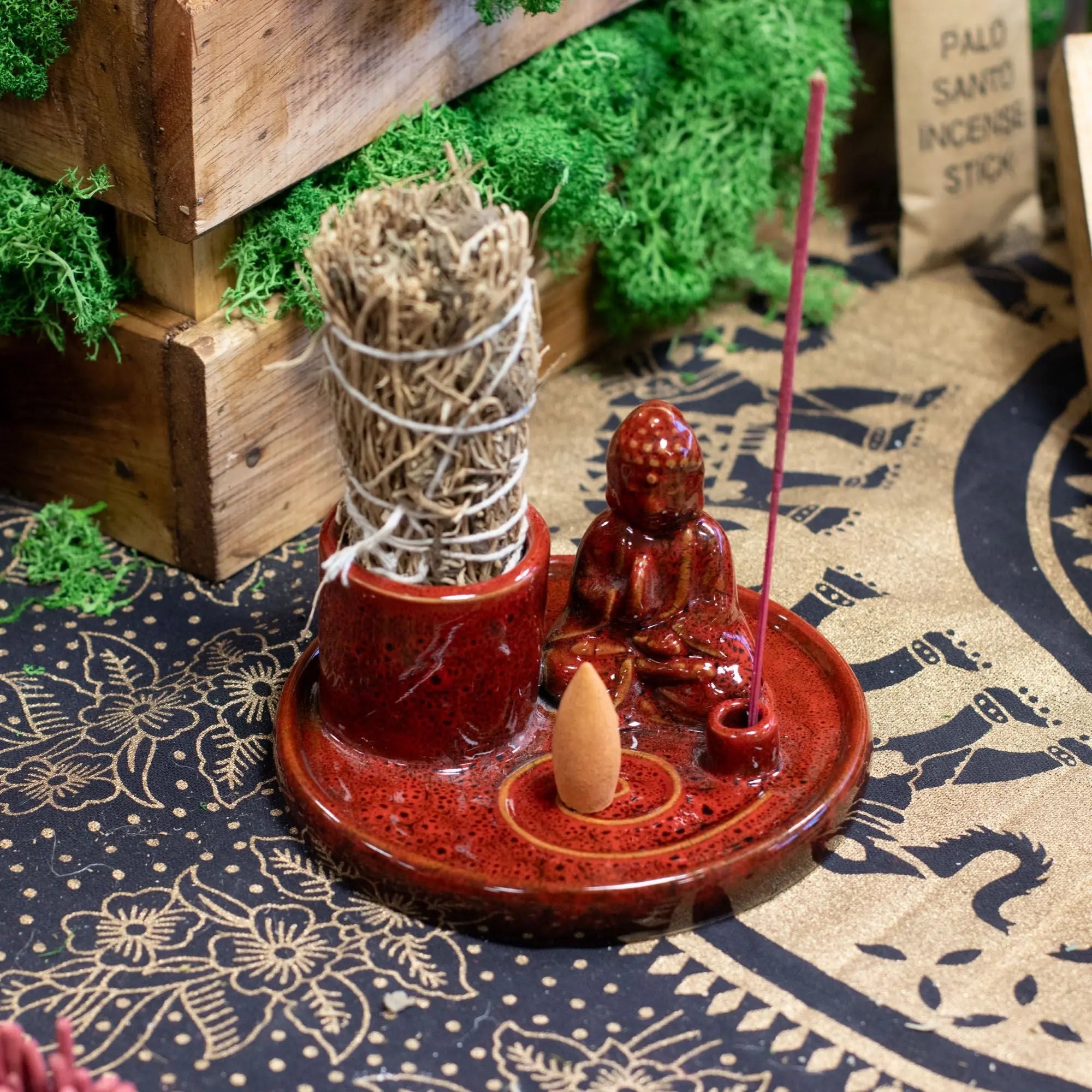 Multi Purpose Buddha Incense Plate - Ceramic Soft Red holding a small Buddha figure, incense cone, stick, and a bundle of Palo Santo on a decorative cloth background.