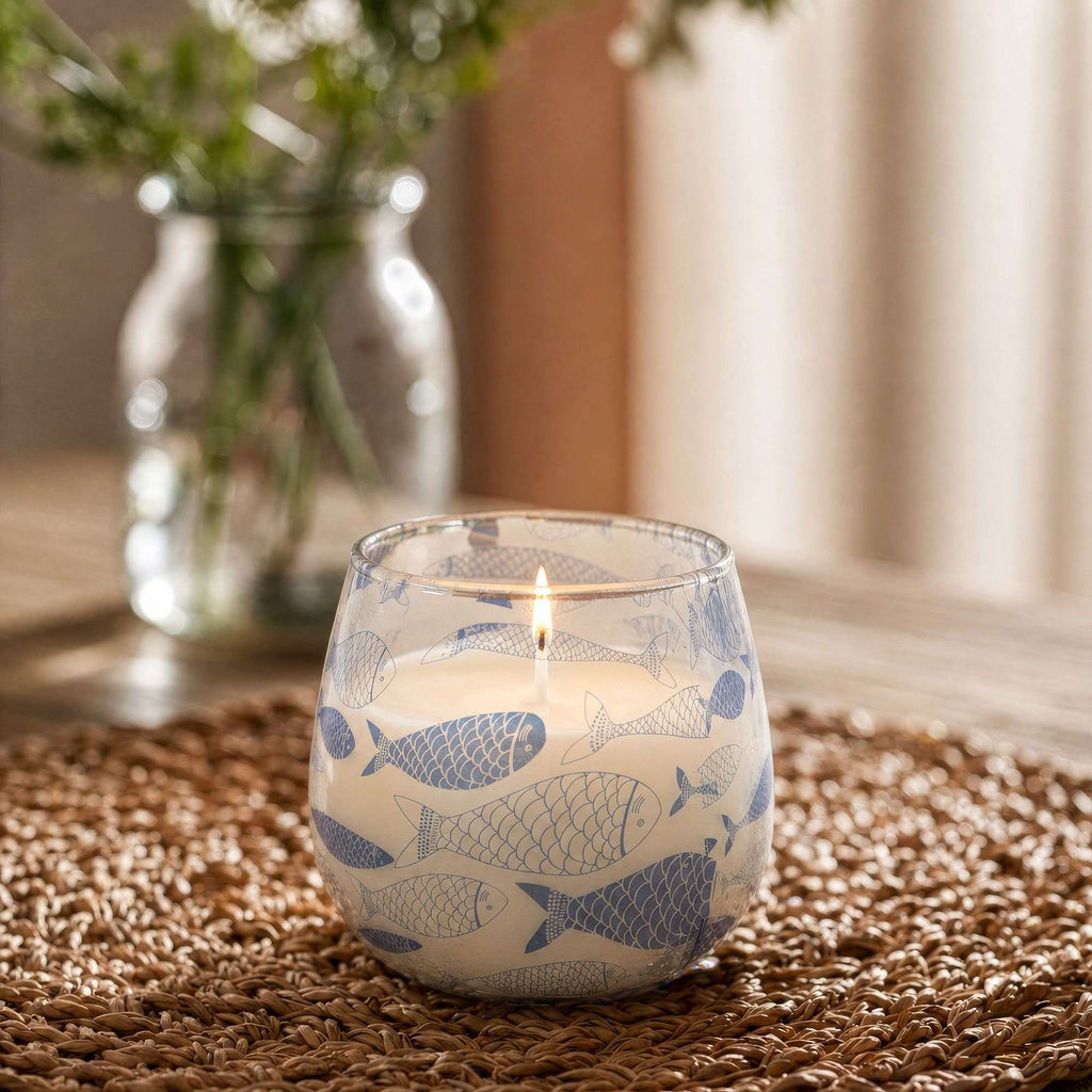 Small Glass Jar Candles - Seabreeze featuring a lit candle inside a glass jar decorated with blue fish patterns, placed on a woven mat with a blurred plant in the background.