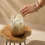 Hand lighting a Tall Glass Jar Candles - Wildlight Woods with dried floral details inside a frosted glass jar placed on a natural wooden table.