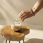 Hand lighting a Small Glass Jar Candles - Drawn by Flame with a matchstick on a natural wooden table, showcasing delicate floral patterns on the glass jar.