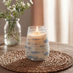 Tall Glass Jar Candles - Seabreeze showing a lit candle in a clear glass jar decorated with blue fish designs, placed on a woven mat with a blurred background of a flower vase and soft lighting.
