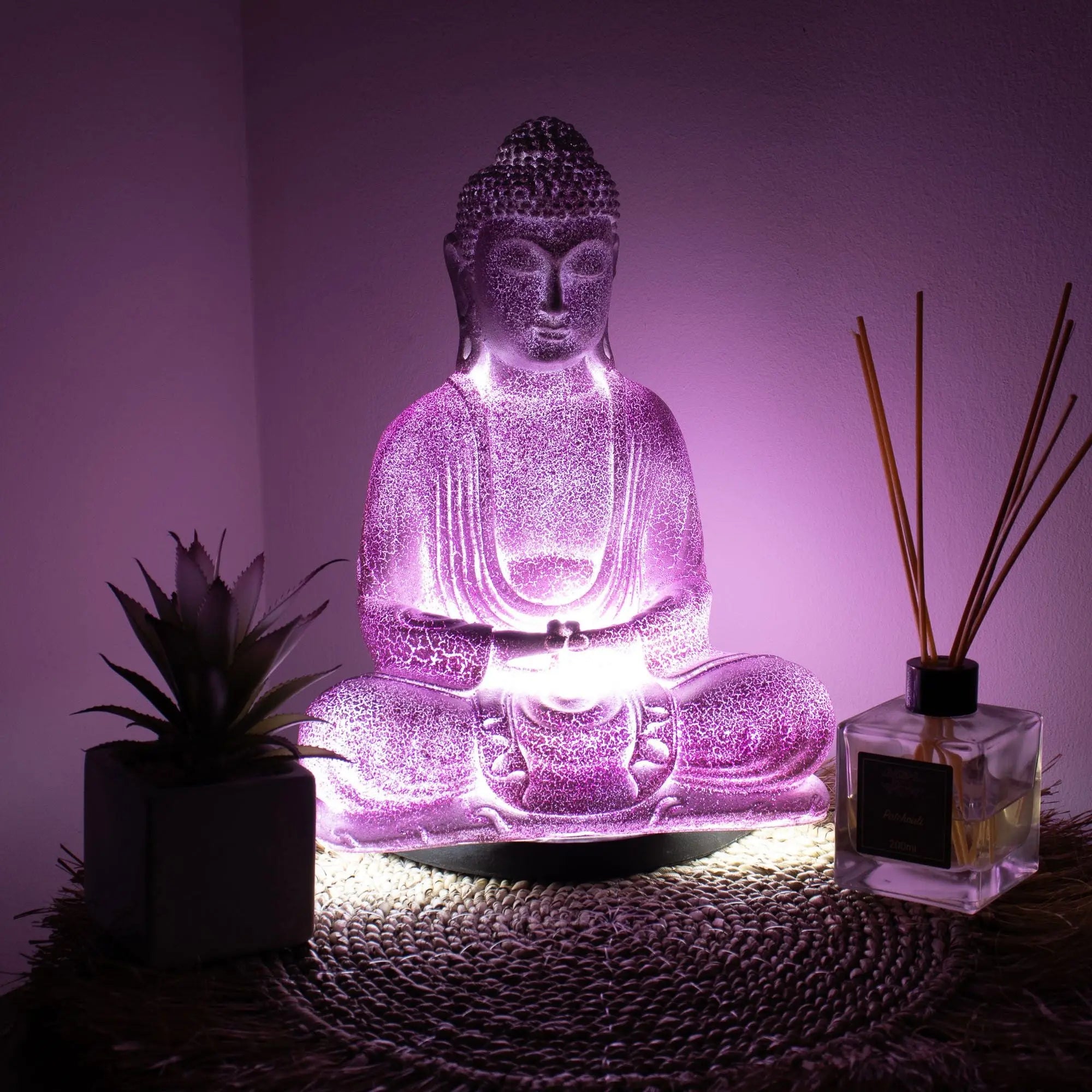 Meditation Buddha Lamp Lavender Crackle - Oval Stand glowing softly in purple light, placed on a woven mat with a small plant and reed diffuser nearby creating a calming decor scene.