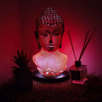 Buddha Bust Lamp Molton Lava - Oval stand glowing with warm red light, placed on a round woven mat next to a plant and reed diffuser, creating a peaceful ambiance.